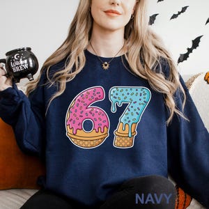May include: Navy blue sweatshirt featuring a colorful graphic of the number 67 designed as a donut and ice cream cone. The number 6 is pink with sprinkles, and the 7 is blue with sprinkles. The word "NAVY" is printed at the bottom.