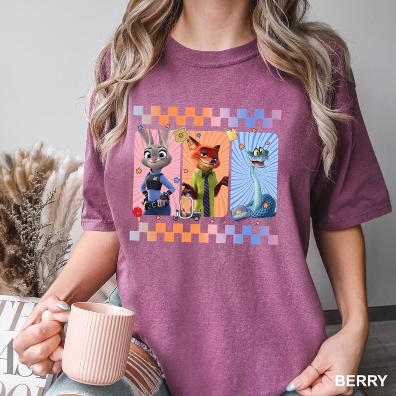 May include: A berry-colored short-sleeve t-shirt featuring a graphic print of three animated characters: a rabbit in a police uniform, a fox in a green shirt and tie, and a blue snake. The design includes a checkered border and small star icons.