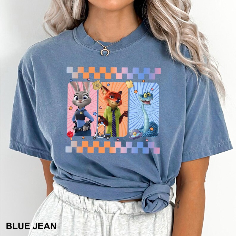 May include: A blue jean colored t-shirt featuring a graphic print of three animated characters from Zootopia, including a rabbit, a fox, and a snake, framed by orange and blue checkered borders and small star icons on a light blue background.