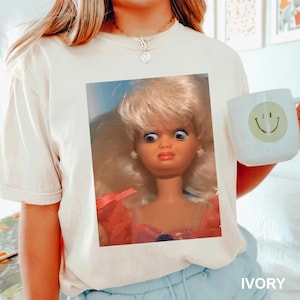 May include: Ivory-colored t-shirt featuring a doll graphic with a surprised expression. The doll has blonde hair, blue eyes, and red lipstick. A white mug with a smiley face is held in the image. The word "IVORY" is printed at the bottom.