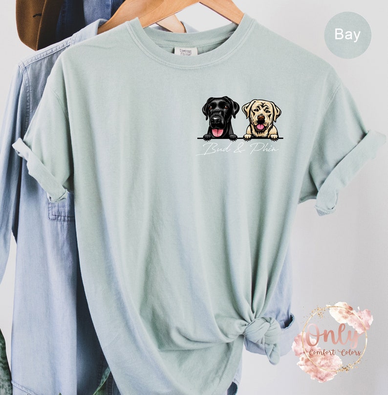 May include: A light blue t-shirt featuring a graphic of two Labrador Retrievers looking over a surface. The text "Bud & Phin" is printed below the dogs.
