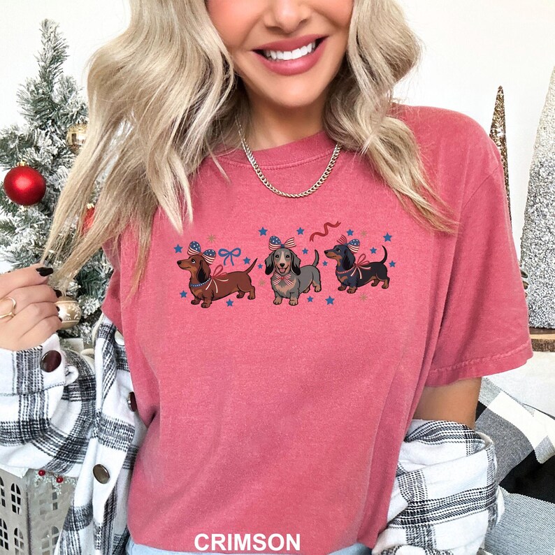 Peut inclure: T-shirt couleur pourpre avec trois teckels portant des n&oelig;uds papillon patriotiques et des &eacute;toiles. Les chiens sont marron, gris et noirs. Le mot "CRIMSON" est imprim&eacute; en bas du t-shirt.