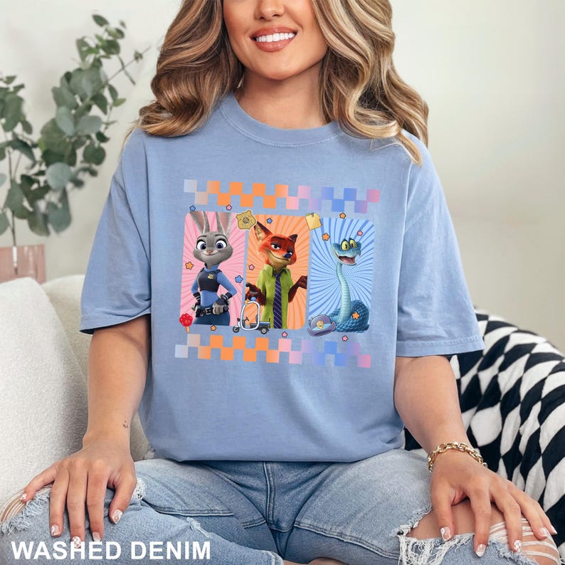May include: A light blue t-shirt featuring a graphic print of three animated characters: a rabbit in a police uniform, a fox in a green shirt and striped tie, and a blue snake. The design is framed by a colorful checkered border.
