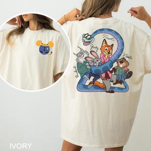 May include: Ivory-coloured t-shirt with a cartoon design on the front and back. The front has a mouse-shaped design with a blue outfit. The back displays a blue snake with cartoon characters, including a fox, rabbit, and beaver. The word "IVORY" is at the bottom.