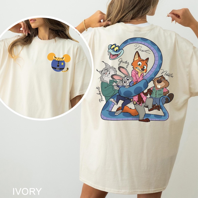 May include: Ivory-coloured t-shirt with a cartoon design on the front and back. The front has a mouse-shaped design with a blue outfit. The back displays a blue snake with cartoon characters, including a fox, rabbit, and beaver. The word "IVORY" is at the bottom.
