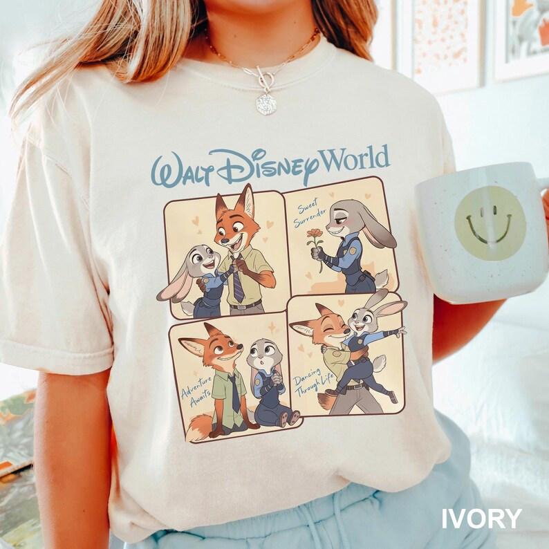 K&ouml;nnte beinhalten: Ein cremefarbenes T-Shirt mit dem Text Walt Disney World und einer viergeteilten grafischen Illustration von Nick Wilde und Judy Hopps aus Zoomania. Die Felder zeigen die Figuren bei Interaktionen mit dem Text Sweet Surrender, Adventure Awaits und Dancing Through Life.