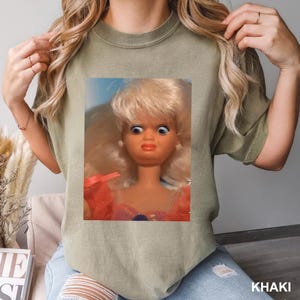 May include: Khaki-colored t-shirt featuring a doll graphic. The doll has blonde hair, blue eyes, and a surprised expression. The word "KHAKI" is printed in white at the bottom right corner of the image.