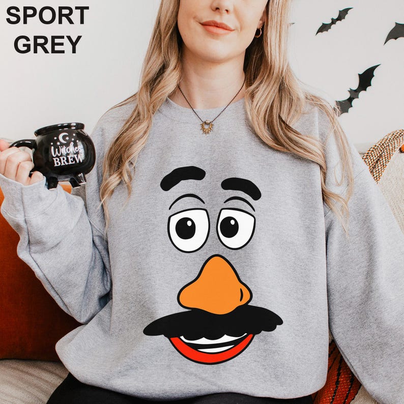 May include: Sport grey sweatshirt featuring a cartoon face with a black mustache, orange nose, and red smile. The sweatshirt has the words "SPORT GREY" printed at the top. A black mug with the words "Witches Brew" is held in the left hand.