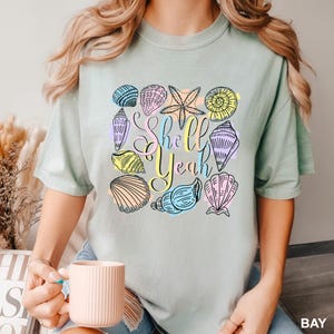 May include: A light green t-shirt featuring a collection of colorful seashell illustrations. The phrase "Shell Yeah" is written in a playful font. The seashells are outlined in black and filled with pastel colors.