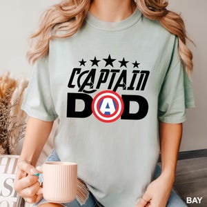 Puede incluir: Camiseta verde claro con la frase "CAPTAIN DAD" en negro, con un escudo rojo, blanco y azul. Encima del texto hay cinco estrellas negras. La camiseta es de estilo casual y de manga corta.