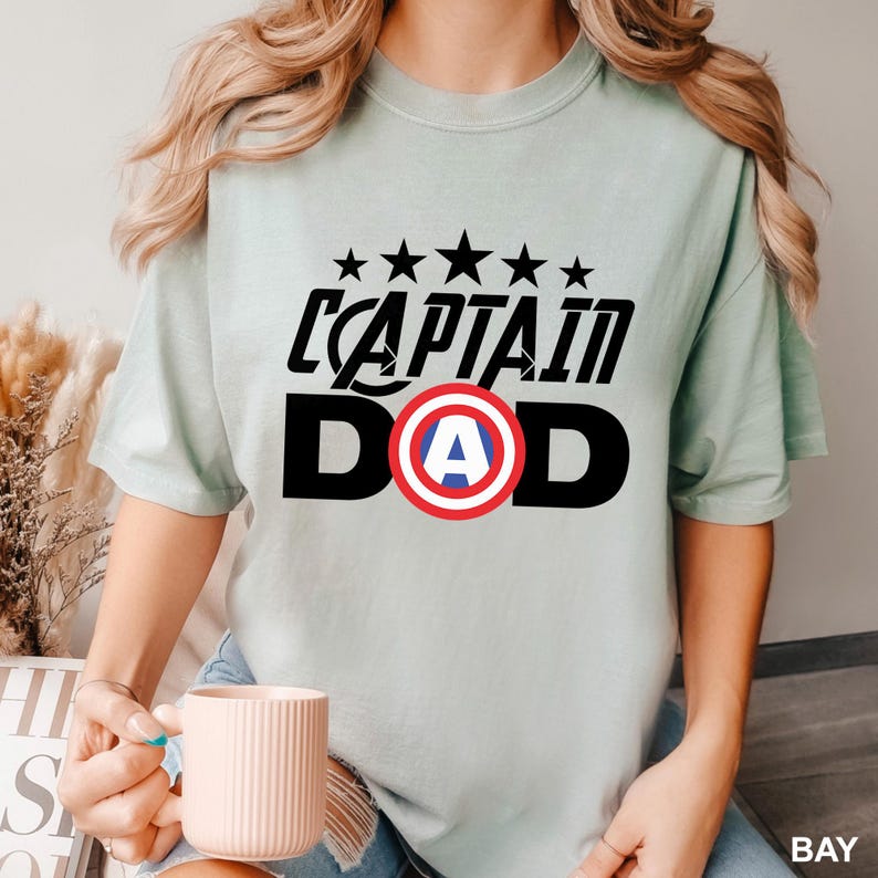 Puede incluir: Camiseta verde claro con la frase "CAPTAIN DAD" en negro, con un escudo rojo, blanco y azul. Encima del texto hay cinco estrellas negras. La camiseta es de estilo casual y de manga corta.
