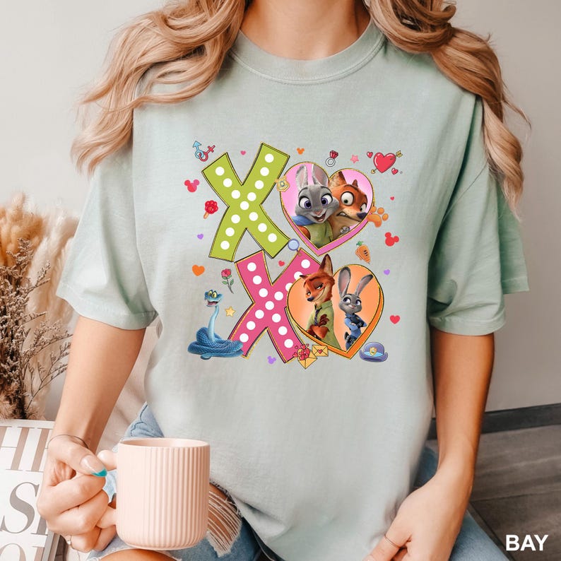 May include: A light green t-shirt featuring the characters from Zootopia. The shirt has a graphic design with the letters "XO" and images of the characters in heart-shaped frames. The word "BAY" is printed in the bottom right corner.