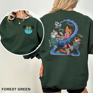 Op de afbeelding: Bostgroen sweatshirt met een cartoonfiguur op de voorkant en een groep cartoonfiguren met een blauwe slang op de achterkant. Het ontwerp aan de voorkant is een blauw gezicht met zwarte ogen en oren. Het ontwerp aan de achterkant bevat de namen "Judy", "Nest" en "Nibbles".