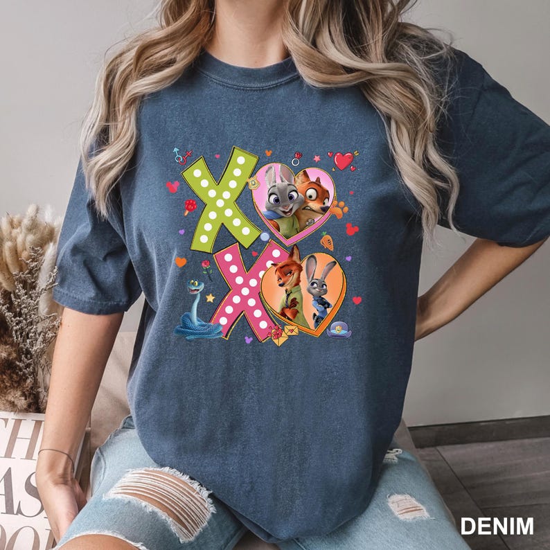 May include: A denim blue t-shirt featuring a colorful graphic design. The design includes the text "XO" with heart-shaped frames containing cartoon characters. The shirt has a crew neck and short sleeves.