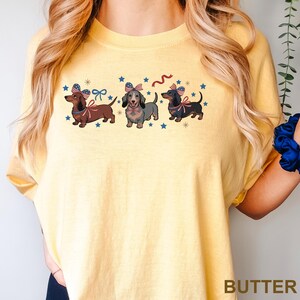 Peut inclure: T-shirt jaune avec trois teckels orn&eacute;s de n&oelig;uds patriotiques et d'&eacute;toiles. Les chiens sont marron, gris et noirs, avec des n&oelig;uds rouges, blancs et bleus. Le mot "BUTTER" est imprim&eacute; en bas du t-shirt.
