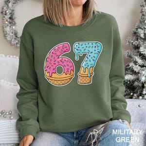 May include: A military green sweatshirt featuring a colorful graphic of the number 67 designed as a donut and ice cream cone. The number 6 is a pink donut with sprinkles, and the 7 is blue ice cream with sprinkles.