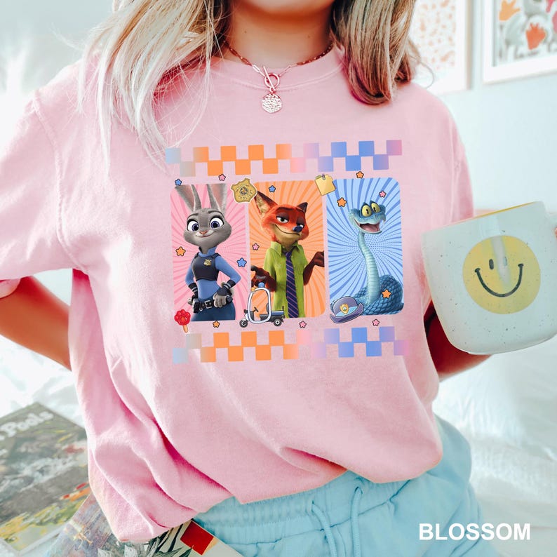May include: A light pink t-shirt featuring a graphic print of three animated characters: a rabbit in a police uniform, a fox in a green shirt and tie, and a blue snake. The design includes a checkered border and small star icons.
