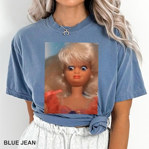 May include: A blue jean colored t-shirt featuring a close-up of a vintage doll with wide eyes and a surprised expression. The text "BLUE JEAN" is printed at the bottom of the shirt.
