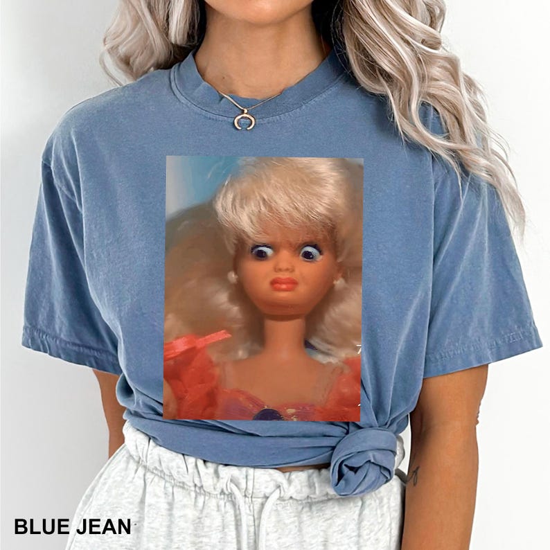 May include: A blue jean colored t-shirt featuring a close-up of a vintage doll with wide eyes and a surprised expression. The text "BLUE JEAN" is printed at the bottom of the shirt.