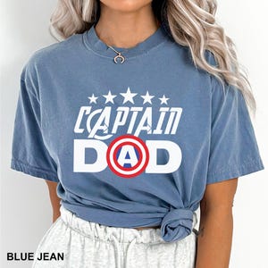 Puede incluir: Camiseta azul vaquero con el texto blanco "CAPTAIN DAD", con un dise&ntilde;o de escudo rojo y azul. La camiseta tiene cuello redondo y mangas cortas. El texto "BLUE JEAN" est&aacute; en la parte inferior de la imagen.