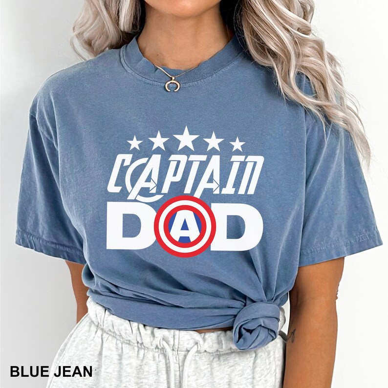 Puede incluir: Camiseta azul vaquero con el texto blanco "CAPTAIN DAD", con un dise&ntilde;o de escudo rojo y azul. La camiseta tiene cuello redondo y mangas cortas. El texto "BLUE JEAN" est&aacute; en la parte inferior de la imagen.