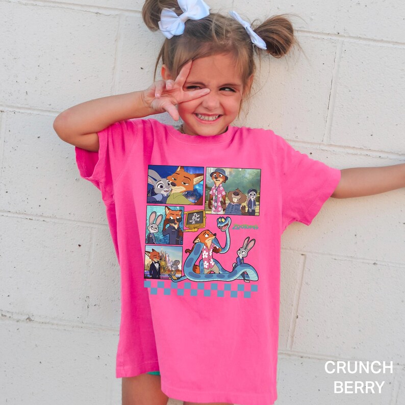 以下が含まれることがあります： 鮮やかなピンク色のTシャツ。カラフルな漫画のデザインで、キツネやウサギなど、さまざまなキャラクターが描かれています。Tシャツの右下には「Crunch Berry」の文字がプリントされています。
