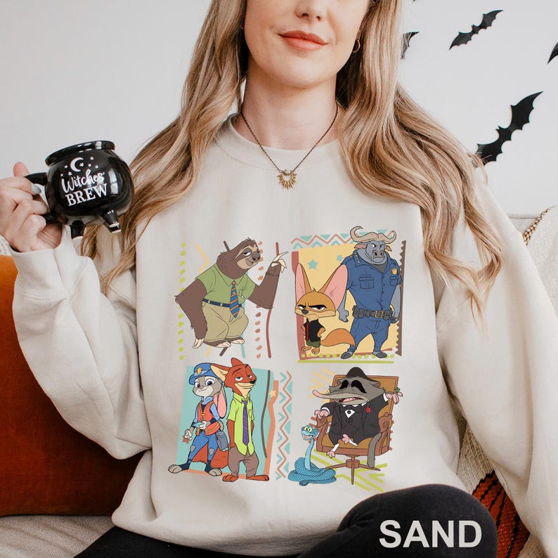 May include: Sand-colored sweatshirt featuring a colorful graphic of characters from the animated movie Zootopia. The design includes images of a sloth, fox, buffalo, rabbit, and other characters. The word "SAND" is printed at the bottom.