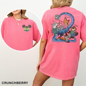 May include: A pink t-shirt with a cartoon design on the front and back. The front has a Mickey Mouse head with a tie. The back has a blue snake with cartoon characters and the names Judy, Nick, and Nibbles. The word "Crunchberry" is at the bottom.