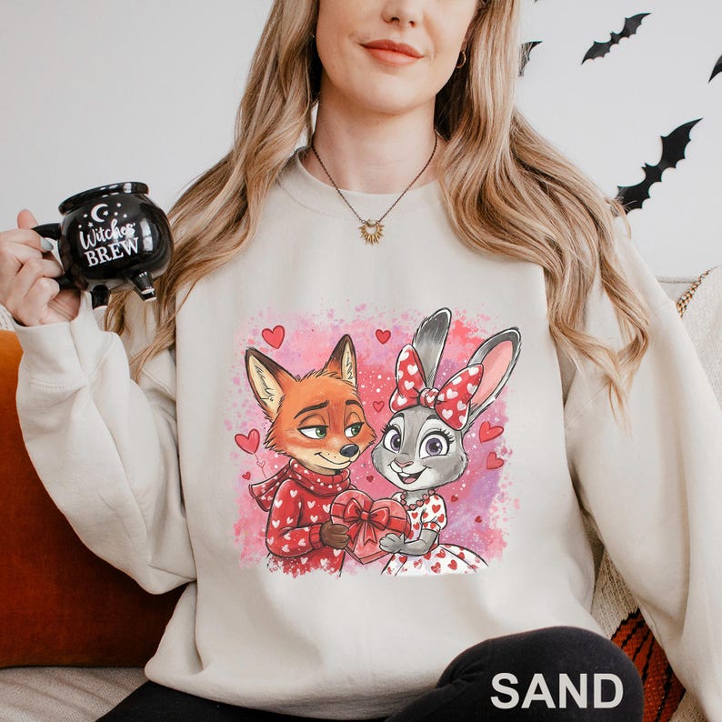 K&ouml;nnte beinhalten: Sandfarbenes Sweatshirt mit einem Cartoon-Fuchs und -Hasen, die ein herzf&ouml;rmiges Geschenk halten. Der Fuchs tr&auml;gt einen roten Schal und Pullover, w&auml;hrend der Hase eine Schleife und ein Kleid mit Herzmustern hat. Rosa Herzen und ein rosa Hintergrund vervollst&auml;ndigen das Design.