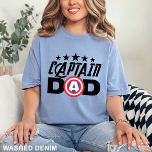 Puede incluir: Camiseta azul claro con la frase "CAPTAIN DAD" en negro y un gr&aacute;fico del escudo del Capit&aacute;n Am&eacute;rica. Encima del texto hay cinco estrellas negras. La camiseta es de un material suave.