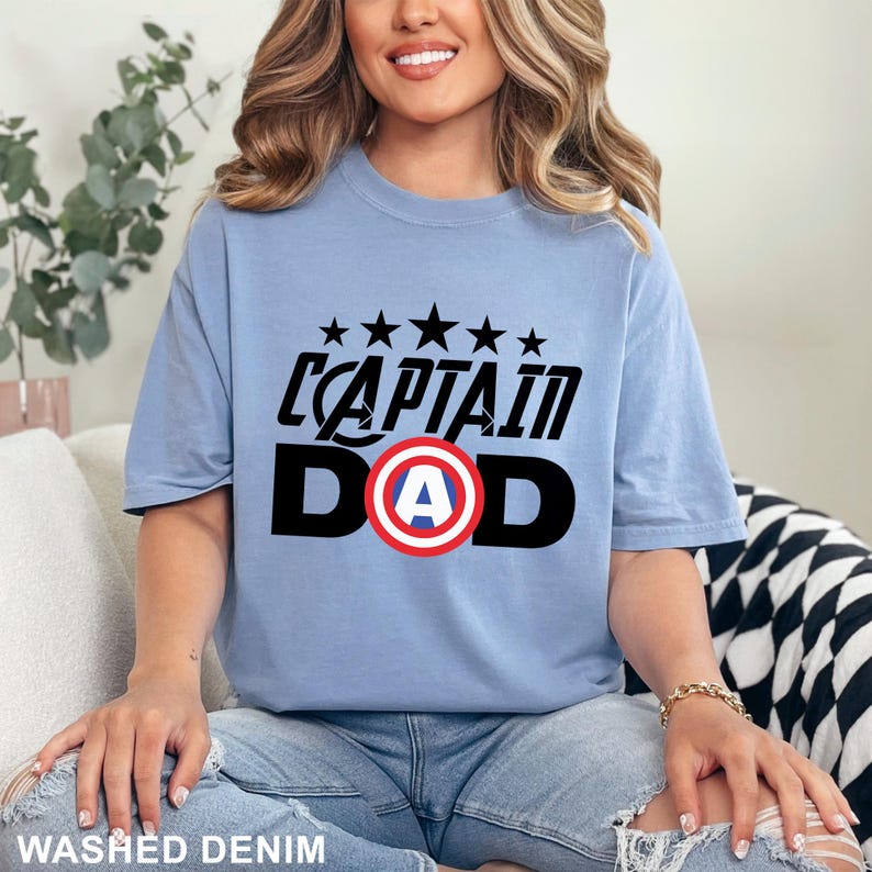 Puede incluir: Camiseta azul claro con la frase "CAPTAIN DAD" en negro y un gr&aacute;fico del escudo del Capit&aacute;n Am&eacute;rica. Encima del texto hay cinco estrellas negras. La camiseta es de un material suave.