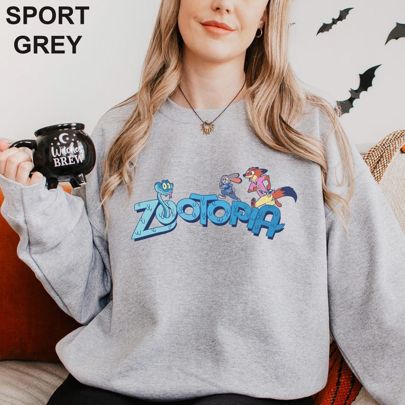 Puede incluir: Una sudadera gris claro con la palabra "ZOOTOPIA" en azul, con personajes de dibujos animados. La sudadera tambi&eacute;n tiene las palabras "SPORT GREY" en la parte superior. Una taza negra con las palabras "Witches Brew" se sostiene en la mano izquierda.