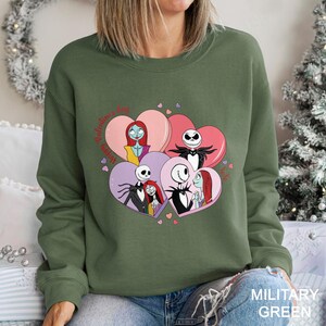 May include: Military green sweatshirt featuring a graphic of characters from The Nightmare Before Christmas inside heart shapes. The design includes Jack Skellington, Sally, and other characters. The words "Happy Valentine's Day" are written in a cursive font.
