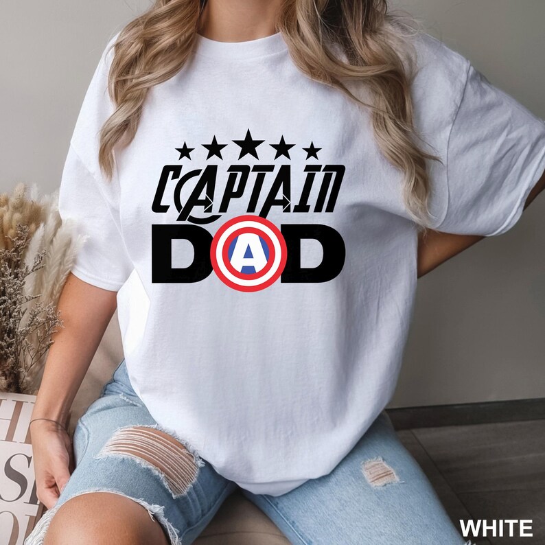 Puede incluir: Camiseta blanca con el texto negro "CAPTAIN DAD", con un escudo rojo, blanco y azul. Encima del texto hay cinco estrellas negras. La camiseta es de estilo casual.