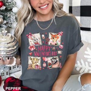 May include: Dark gray t-shirt with a Valentine's Day theme. The shirt features four framed cartoon characters, hearts, gifts, and the text "HAPPY VALENTINE'S DAY" in pink. The shirt is a comfortable, casual style.