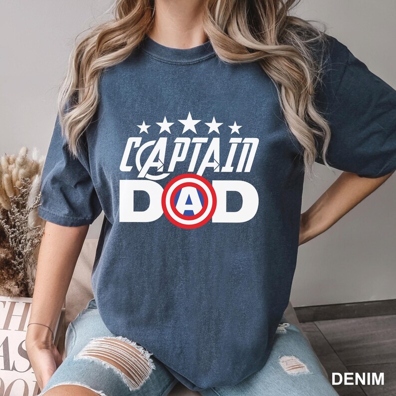 Puede incluir: Una camiseta azul denim con la inscripci&oacute;n blanca "CAPTAIN DAD", con un dise&ntilde;o de escudo. La camiseta tiene cuello redondo y mangas cortas. La palabra "DENIM" est&aacute; impresa en la esquina inferior derecha.