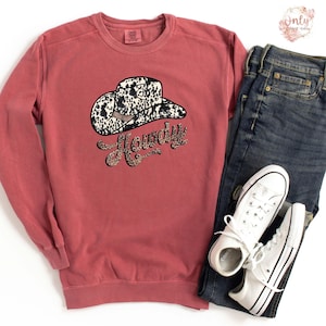 May include: A maroon sweatshirt with a white and black cow print cowboy hat and the word "Howdy" in a black and white cow print font.