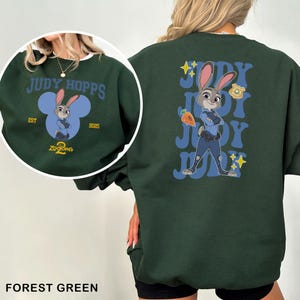 K&ouml;nnte beinhalten: Forstgr&uuml;nes Sweatshirt mit Judy Hopps aus Zootopia. Die Vorderseite hat ein kreisf&ouml;rmiges Design mit Judy Hopps und Text. Die R&uuml;ckseite zeigt Judy Hopps in Polizeiuniform, mit Text und Sterngrafiken. Das Sweatshirt ist ein Rundhals.