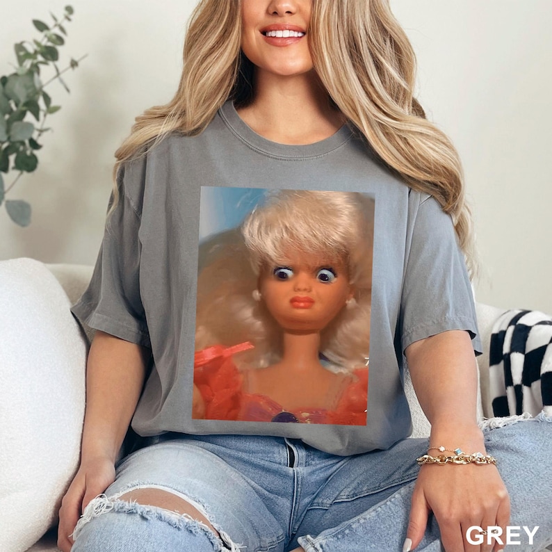 May include: A grey t-shirt featuring a close-up of a vintage doll with wide eyes and a surprised expression. The doll has blonde hair and is wearing a pink and orange dress. The word "GREY" is printed at the bottom right.