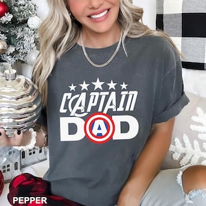Puede incluir: Camiseta gris oscuro con el texto blanco "CAPTAIN DAD", con un dise&ntilde;o de escudo del Capit&aacute;n Am&eacute;rica. Encima del texto hay cinco estrellas blancas. La camiseta es de estilo casual.