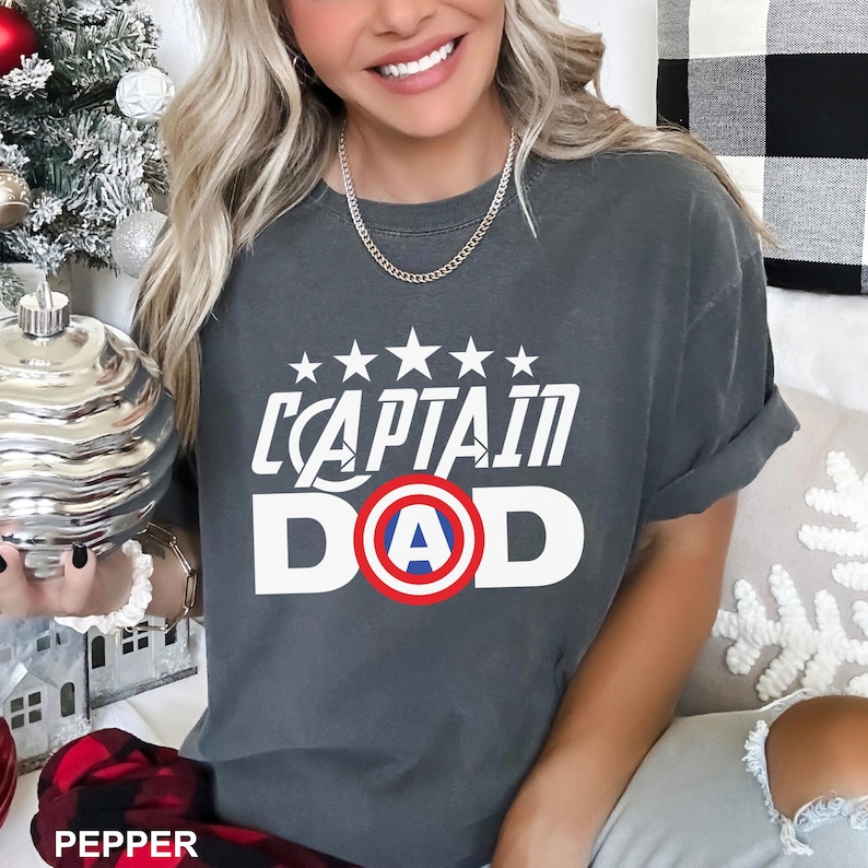 Puede incluir: Camiseta gris oscuro con el texto blanco "CAPTAIN DAD", con un dise&ntilde;o de escudo del Capit&aacute;n Am&eacute;rica. Encima del texto hay cinco estrellas blancas. La camiseta es de estilo casual.