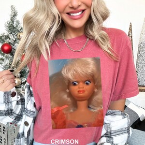 May include: A crimson short-sleeved t-shirt featuring a graphic print of a blonde doll with wide, surprised eyes and an orange dress. The word CRIMSON is printed in white capital letters at the bottom of the shirt.
