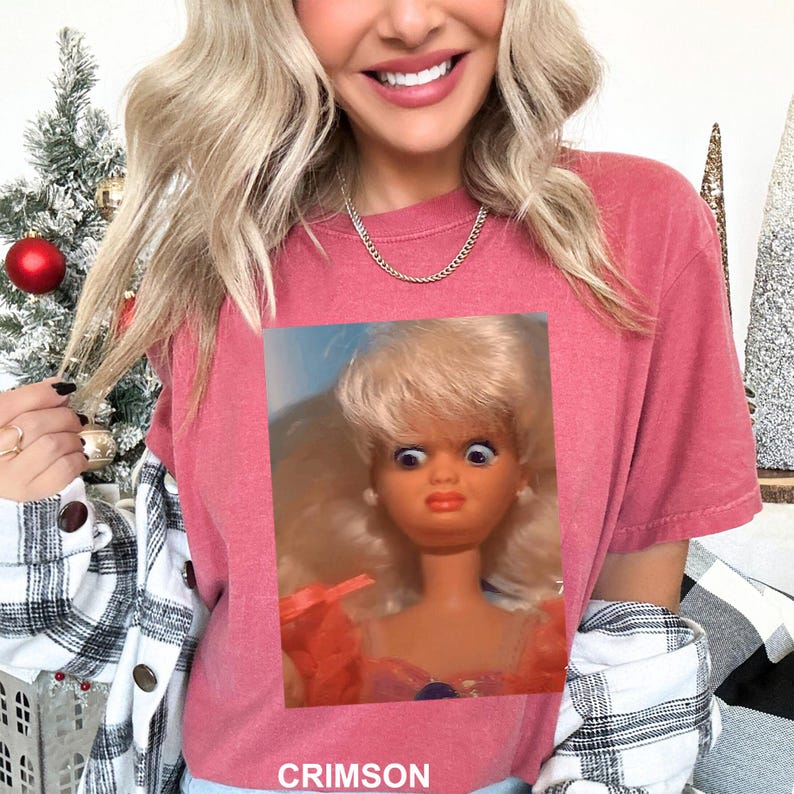 May include: A crimson short-sleeved t-shirt featuring a graphic print of a blonde doll with wide, surprised eyes and an orange dress. The word CRIMSON is printed in white capital letters at the bottom of the shirt.