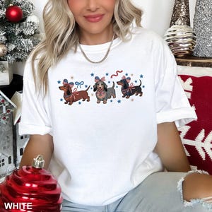 Peut inclure: T-shirt blanc avec un motif de trois teckels portant des n&oelig;uds patriotiques, des &eacute;toiles et des rubans. Le mot "WHITE" est imprim&eacute; en bas de l'image.