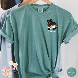 May include: Light green t-shirt with a cartoon image of a calico cat on the front pocket. The cat is looking to the left and the text "Charlie" is written below the cat.