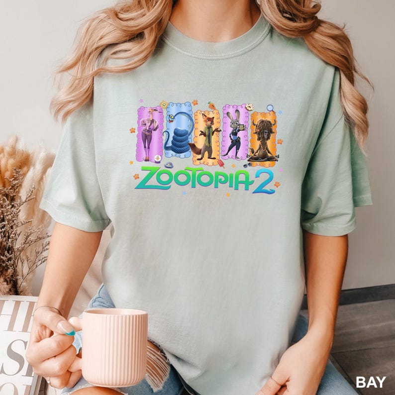 K&ouml;nnte beinhalten: Ein helles salbeigr&uuml;nes T-Shirt mit kurzen &Auml;rmeln und einem Grafikdruck der Zootopia 2-Figuren, darunter eine Giraffe, eine Schlange, ein Fuchs, ein Hase und ein Yak in bunten rechteckigen Rahmen, mit dem Text Zootopia 2 in Blaugr&uuml;n darunter.