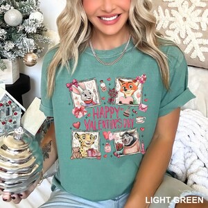 May include: Light green t-shirt with a Valentine's Day graphic. The graphic features cartoon characters in frames, hearts, and the text "Happy Valentine's Day". The shirt is made of a soft material.
