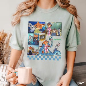 以下が含まれることがあります： 映画「ズートピア」のキャラクターが登場するコミックストリップデザインのライトグリーンのTシャツ。デザインには、キツネ、ウサギ、その他のキャラクターがさまざまなシーンで登場します。「ズートピア」の文字は緑色で印刷されています。