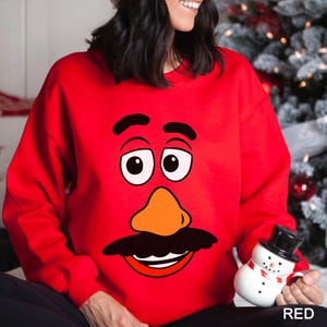 May include: A red sweatshirt featuring a cartoon face design with black eyebrows, eyes, mustache, and an orange nose. The word "RED" is printed in the bottom right corner. The sweatshirt is a festive holiday item.