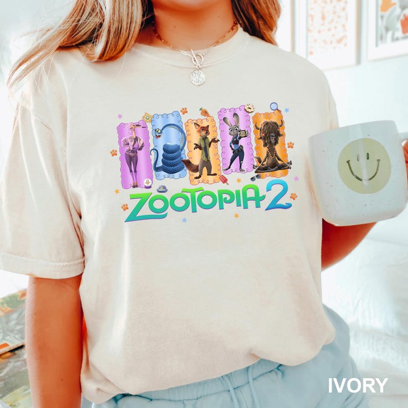 K&ouml;nnte beinhalten: Ein elfenbeinfarbenes Kurzarm-T-Shirt mit einem Grafikdruck von f&uuml;nf Zootopia-Figuren in bunten, gewellten Rechtecken &uuml;ber dem Text Zootopia 2 in Blaugr&uuml;n. Das T-Shirt wird mit einer wei&szlig;en Tasse mit einem gelben Smiley-Gesicht kombiniert.